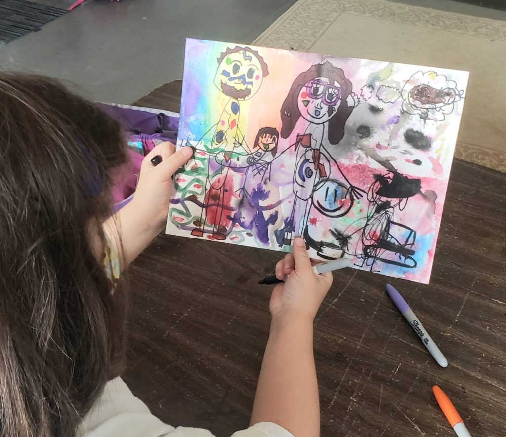 An image from over the shoulder of a child holding and looking at a drawing of her family.
