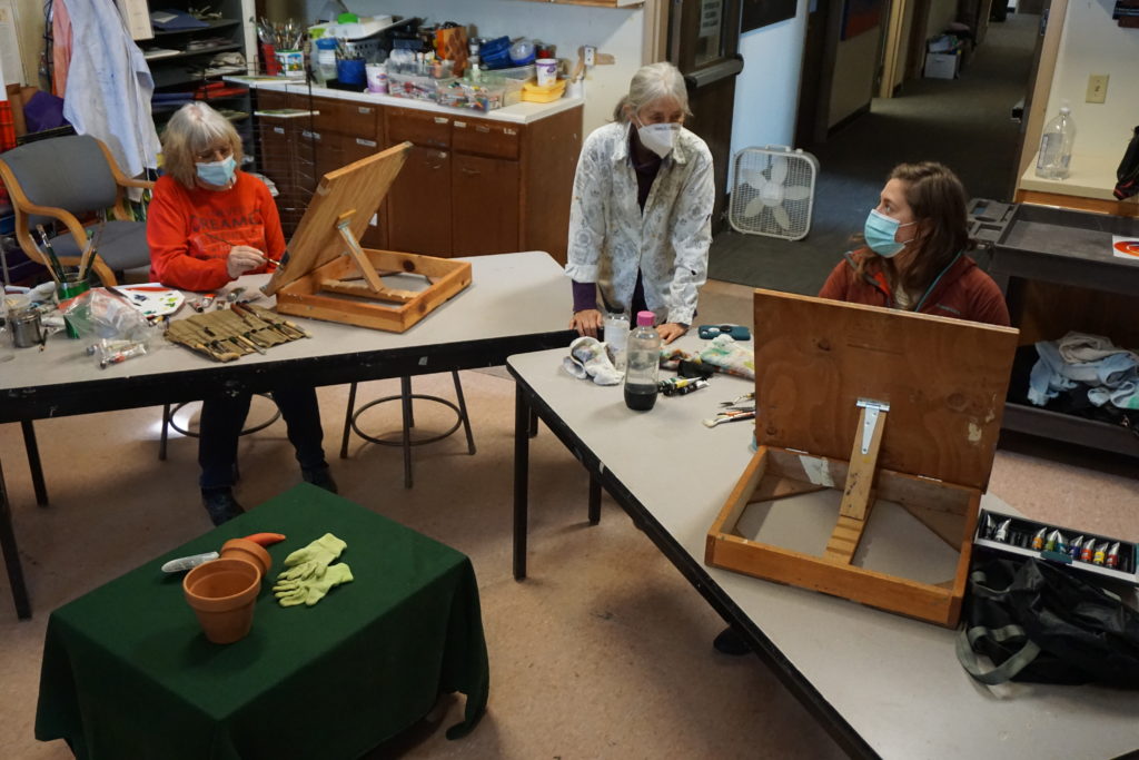 A view of the painting classroom. Two students with easels are painting a still life set-up while the instructor observes.